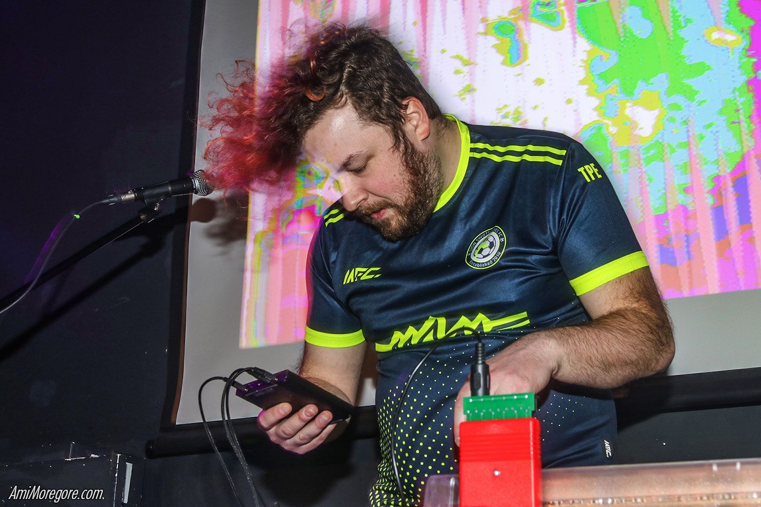 An image of performer DonutShoes currently engaged in a show. He stands holding a device in his hand with a microphone to his left and a screen casting vibrant visuals behind.