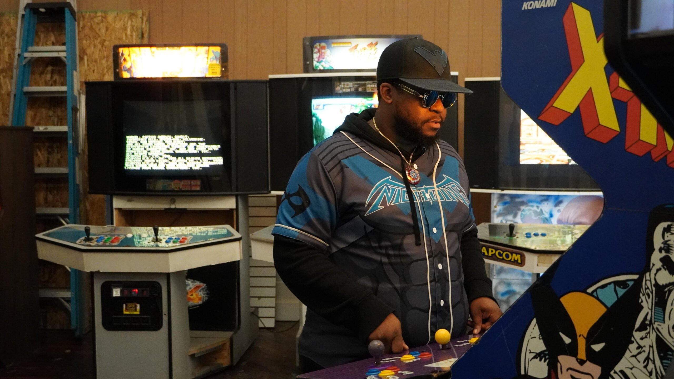 A photo of announced performer, GR3YS0N. He stands in front of an X-Men arcade machine and surrounded by other cabinets in the background.