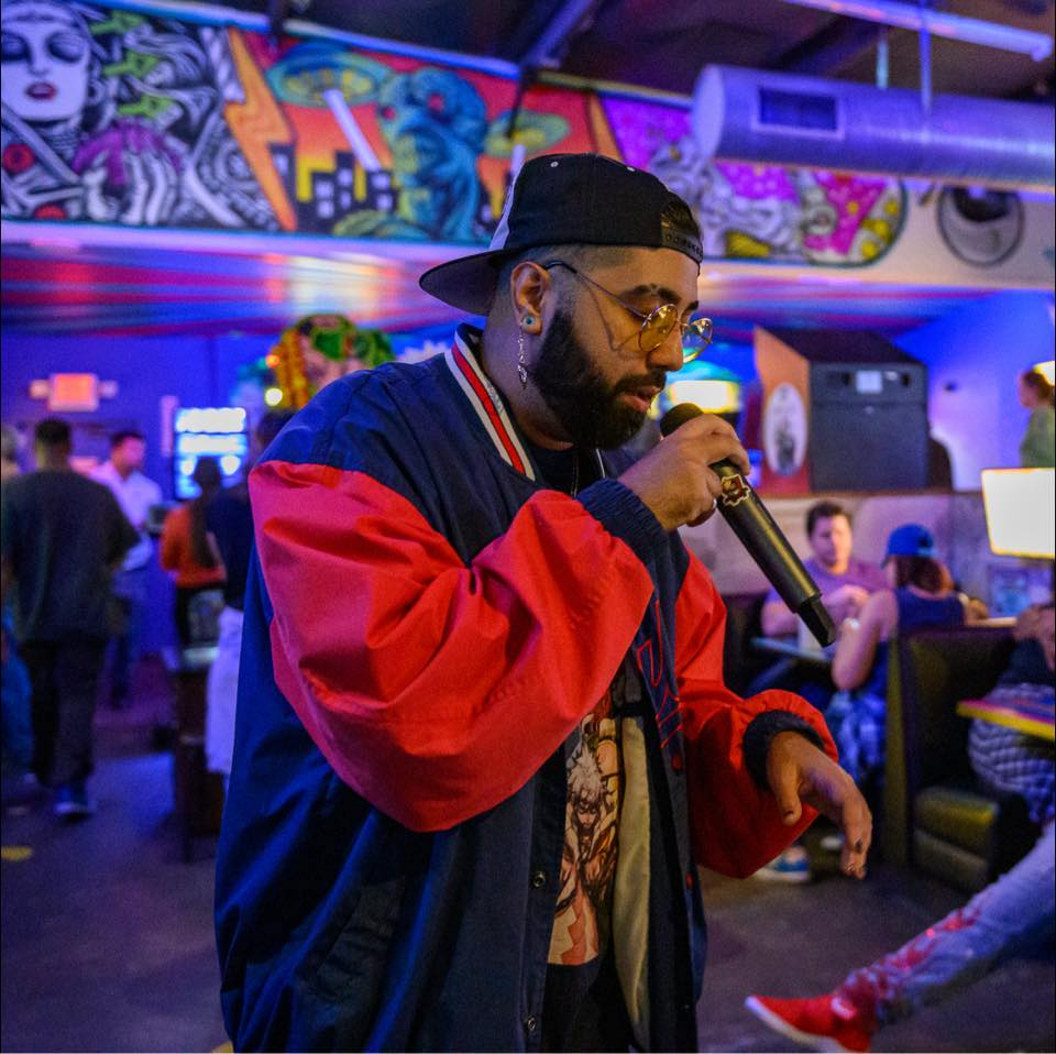 A photo of featured performer, Lord Xzae. He stands amidst a room with a scattered crowd. Some seated, others walking. Xzae is mid performance with a mic in his hand and a focus on lyrical delivery.