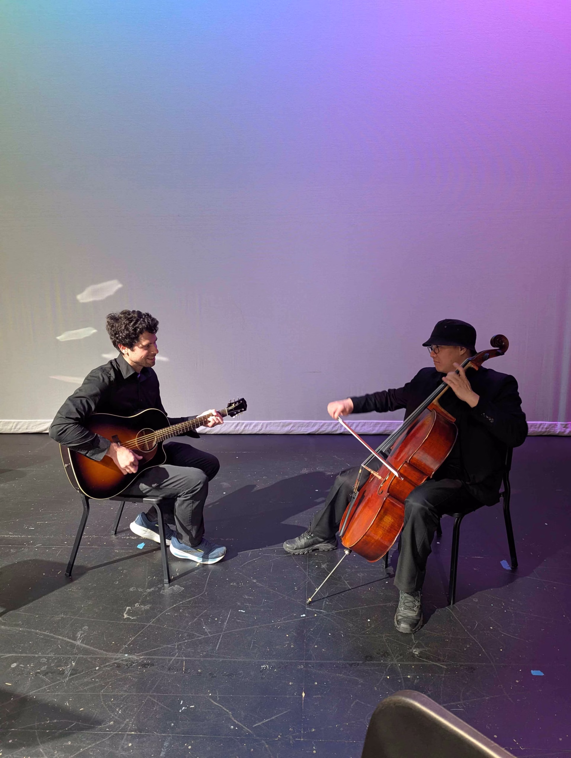 A photo of 7:30 Riders is displayed. The duo sits on a stage with a gentle, blank backdrop; soft cool lighting behind them, and facing each other while performing. A cello and acoustic guitar are featured instruments in this image.