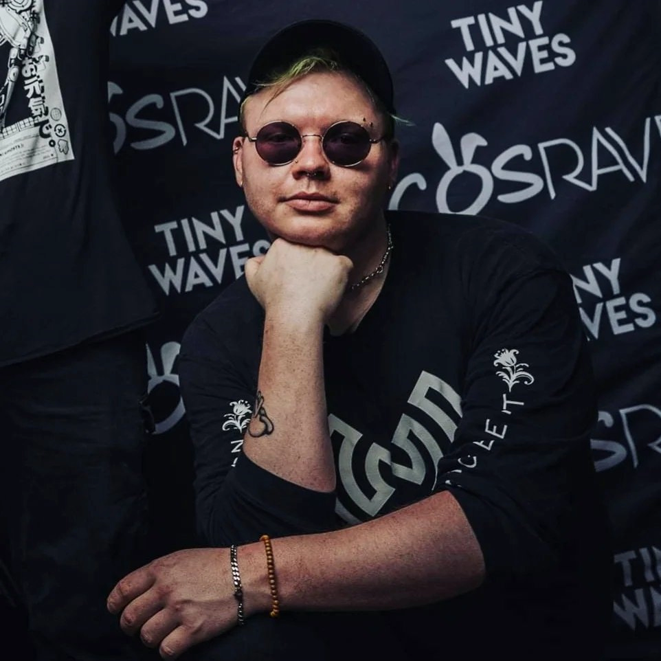 A photo for MAGStock performer Nokbient is displayed. They strike a cool and non-chalant pose, chin resting on their fist with their arms slightly crossed. They smirk behind sunglasses while standing in front of a backdrop featuring Tiny Waves and CosRave branding.